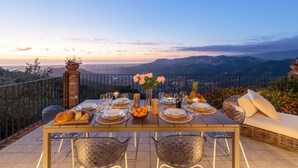 Une table à manger dressée pour quatre personnes, avec une vue panoramique, un canapé en osier et une plante en pot.