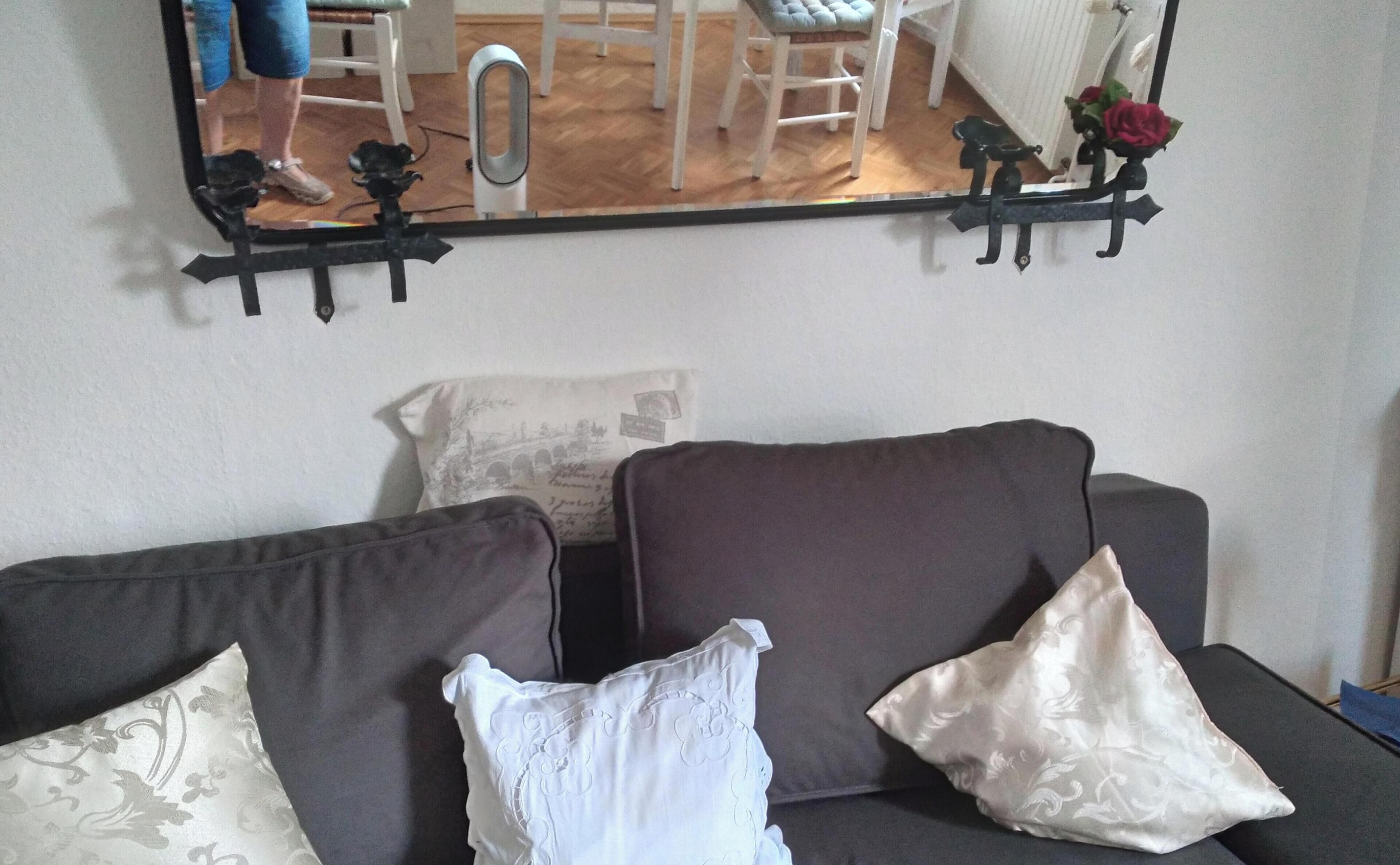 Ein Zimmer mit einem dunkelgrauen Sofa, zwei gemusterten Kissen und einem an der Wand befestigten Spiegel, der einen Teil des Raumes reflektiert.