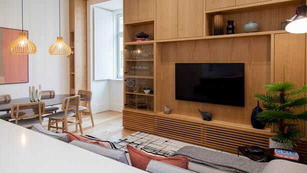 A modern living room with a large flat-screen TV, wooden shelving, and a dining area with chairs and a table.