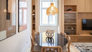 A modern dining area with a round table, chairs, a hanging light fixture, and wooden shelving with glassware.
