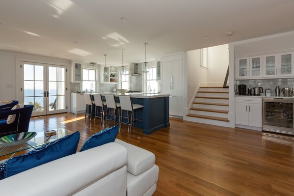 A spacious living area with a white sofa, blue cushions, and a dining table with chairs. A kitchen with white cabinets and a blue island is visible in the background.