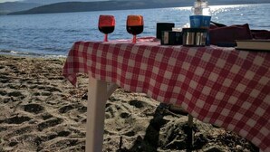 Une table recouverte d’une nappe à carreaux rouges et blancs, avec deux verres à vin et une bouteille d’eau, placée sur une plage d’où l’on aperçoit la mer et des montagnes au loin.