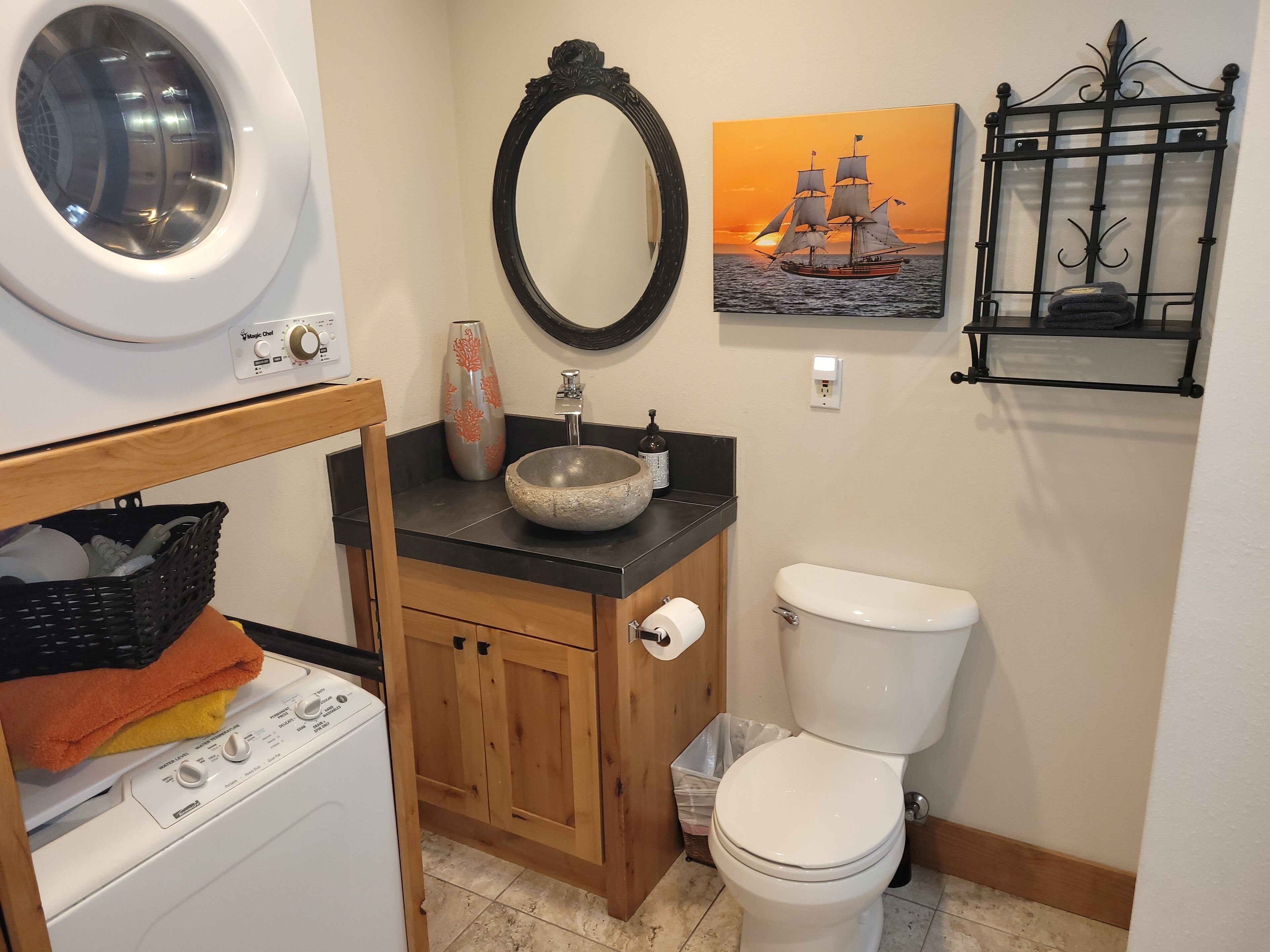 Une petite salle de bain comprenant un lave-linge, un lavabo agrémenté d’un vase décoratif, des toilettes et une étagère fixée au mur.
