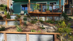 Une maison à l’extérieur verdoyant, dotée d’une terrasse en bois et d’un jardin où poussent différentes plantes, agrémenté d’un petit élément décoratif aquatique.