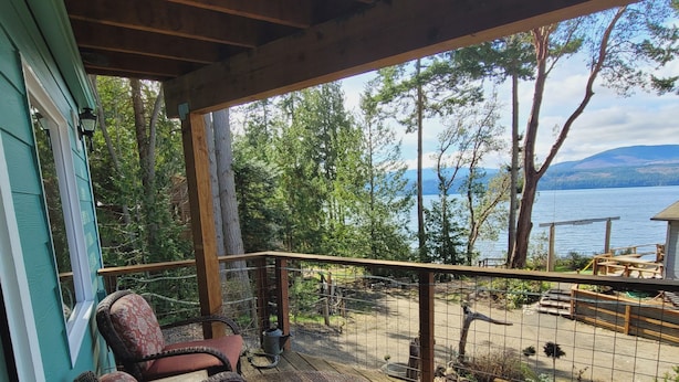 Une terrasse couverte, dotée de chaises en osier et d’une table en verre, qui offre une vue sur un lac et une zone boisée.