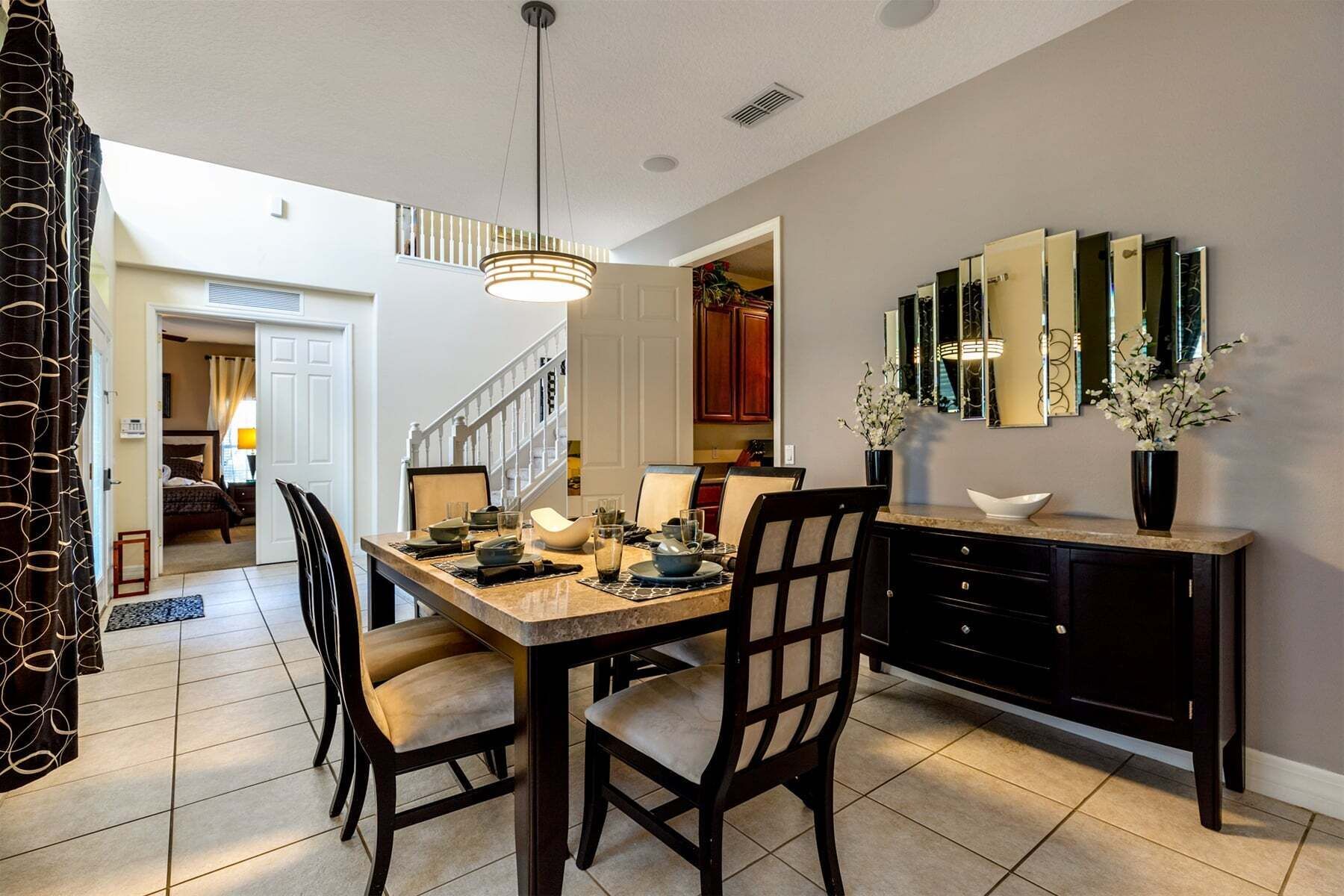 A dining area with a table set for four, chairs, a chandelier, a kitchen in the background, and a staircase.