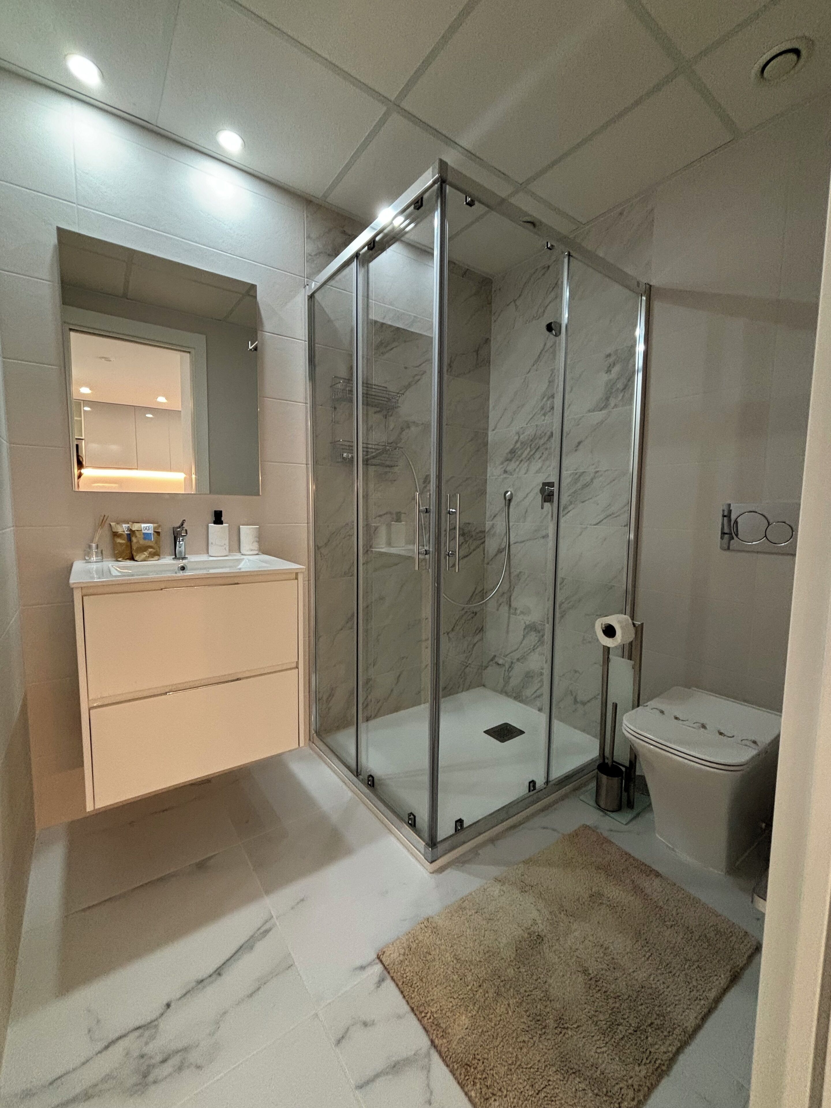 Un baño moderno con ducha de vidrio, inodoro, lavabo con espejo y una alfombra color beige en el piso.