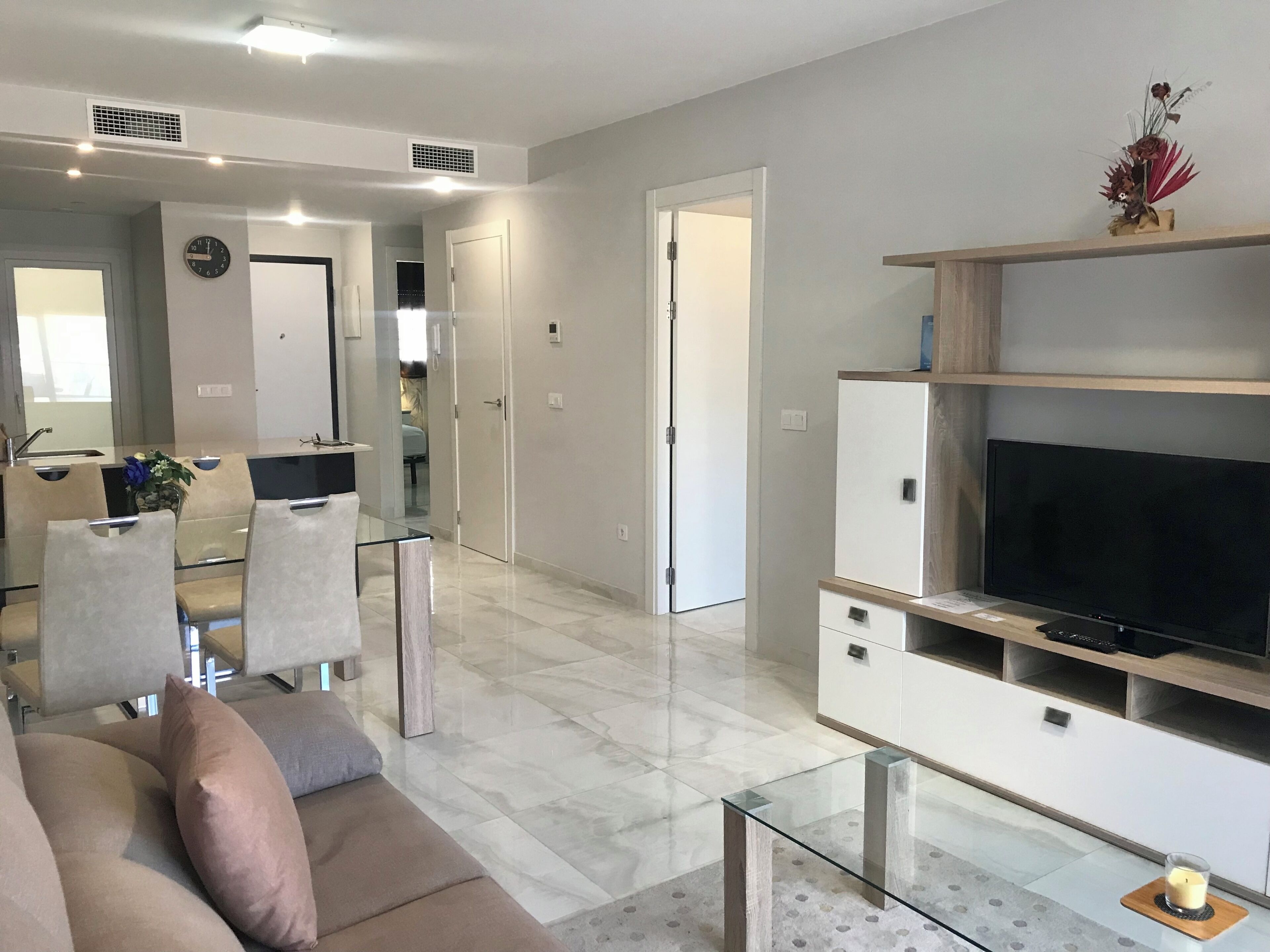 Una sala de estar moderna con sofá, televisión, mesa de centro de vidrio y una cocina al fondo.