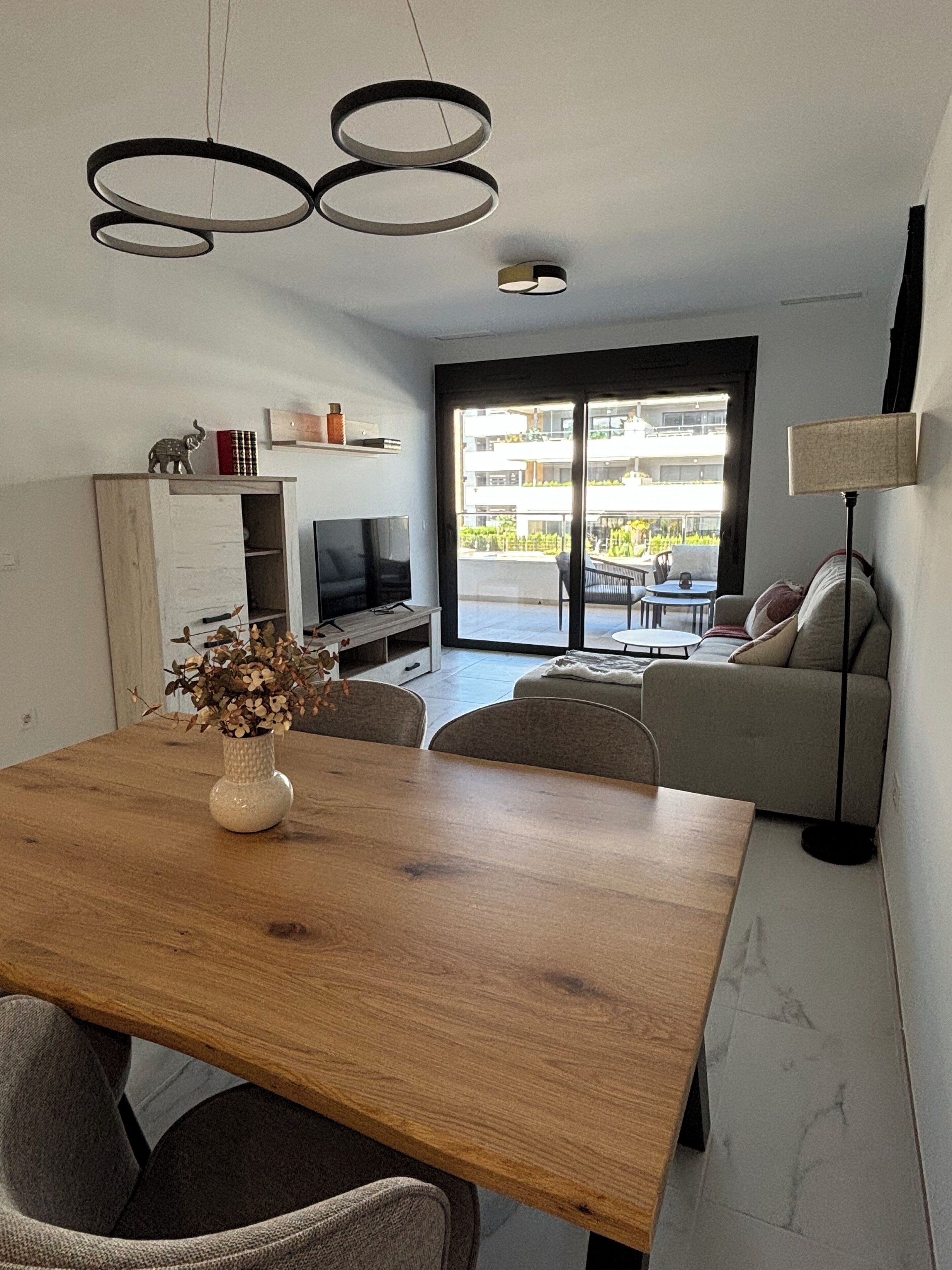 A modern living room with a wooden dining table, a sofa, a TV, and a balcony with a view of buildings.