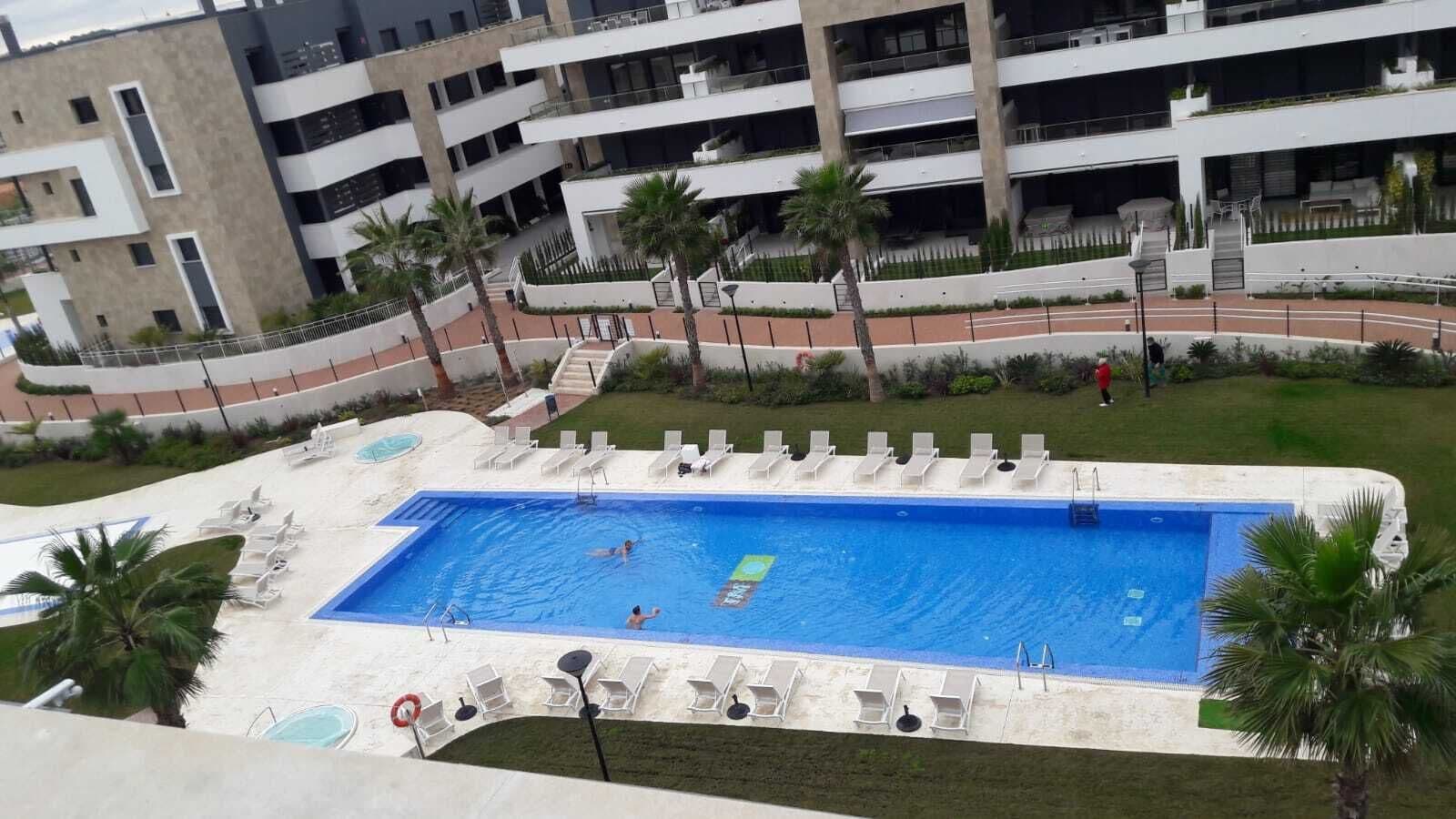 Una piscina con múltiples carriles, rodeada de tumbonas y palmeras, con un edificio de varios pisos al fondo.
