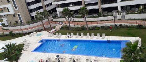 Una piscina con múltiples carriles, rodeada de tumbonas y palmeras, con un edificio de varios pisos al fondo.