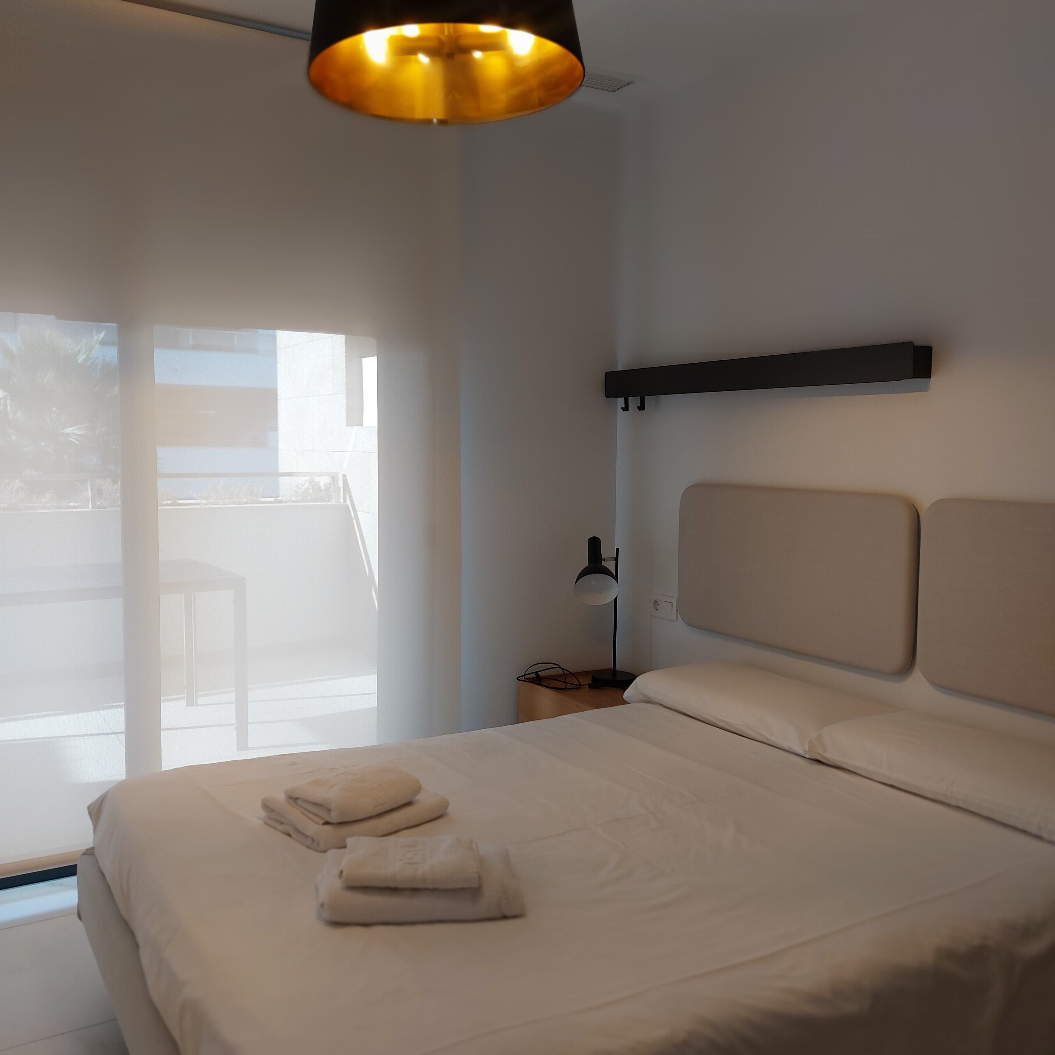 A bedroom with a bed, pillows, a nightstand, a lamp, and a view of the outside.