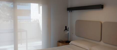 A bedroom with a bed, pillows, a nightstand, a lamp, and a view of the outside.