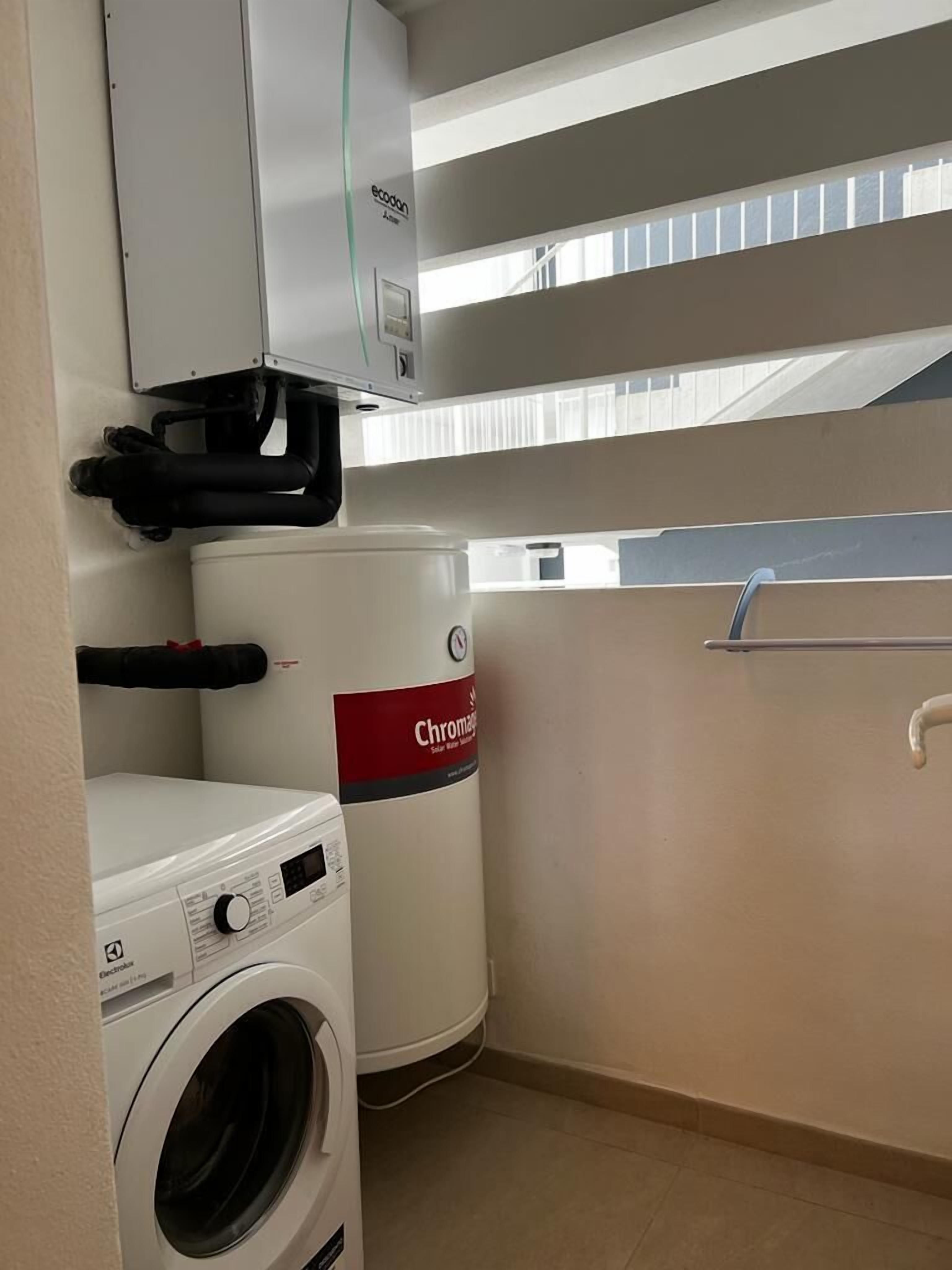 A compact laundry room with a washing machine, a large water heater, and a window with blinds.