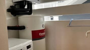 A compact laundry room with a washing machine, a large water heater, and a window with blinds.