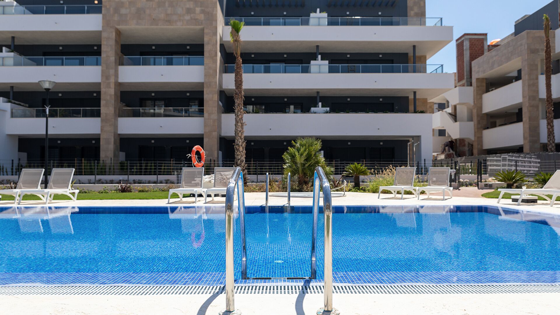A swimming pool with a lifebuoy, surrounded by lounge chairs and palm trees, with a multi-story building in the background.