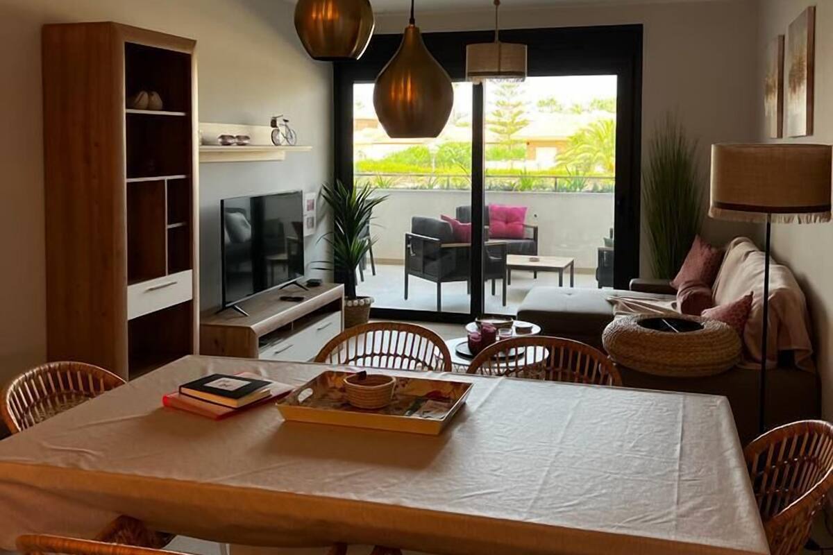 A modern living room with a dining area, featuring a large table, wicker chairs, a TV, and a balcony with outdoor furniture.
