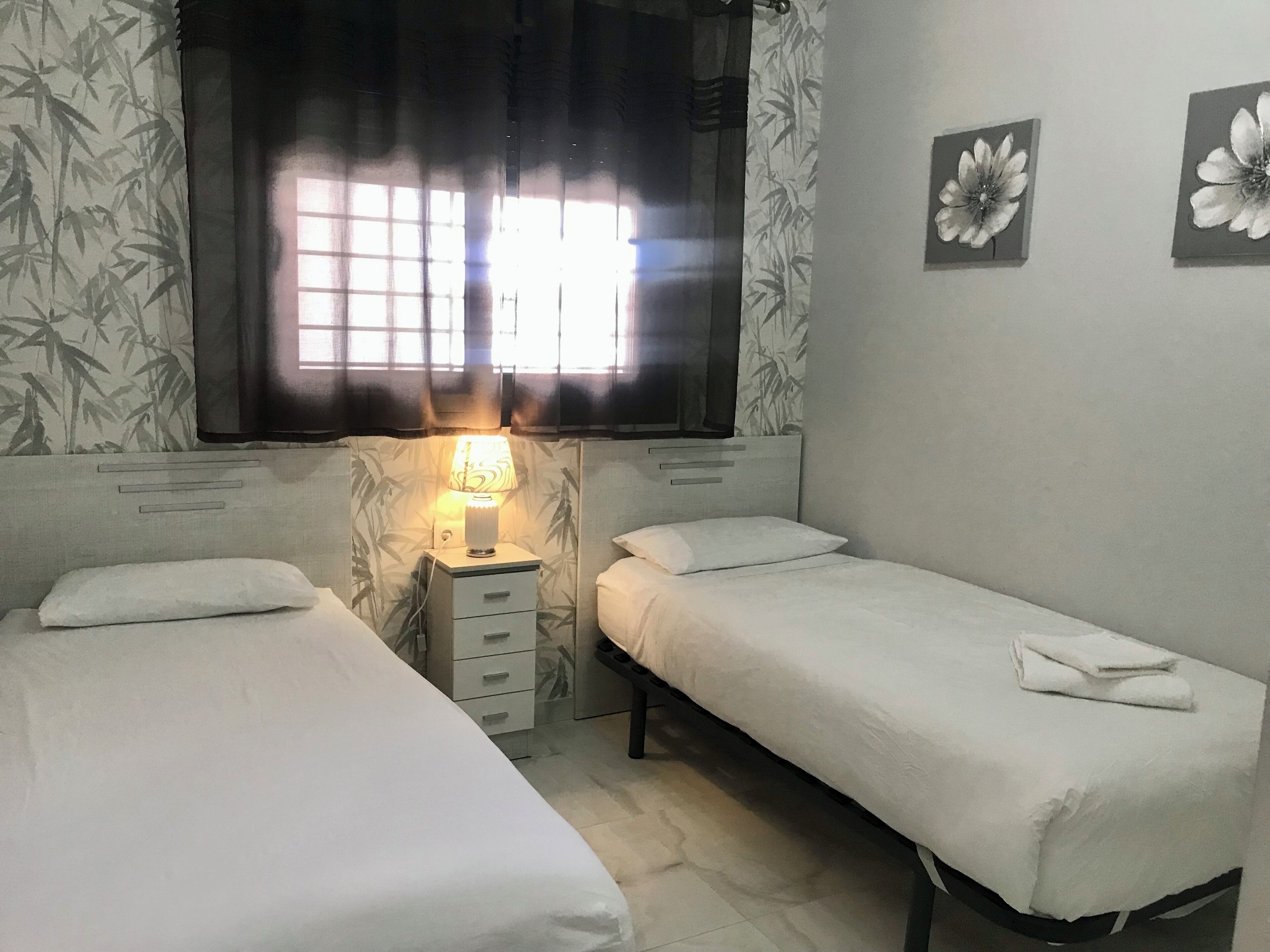 A room with two single beds, a bedside table with a lamp, a window with curtains, and two wall-mounted flower decorations.