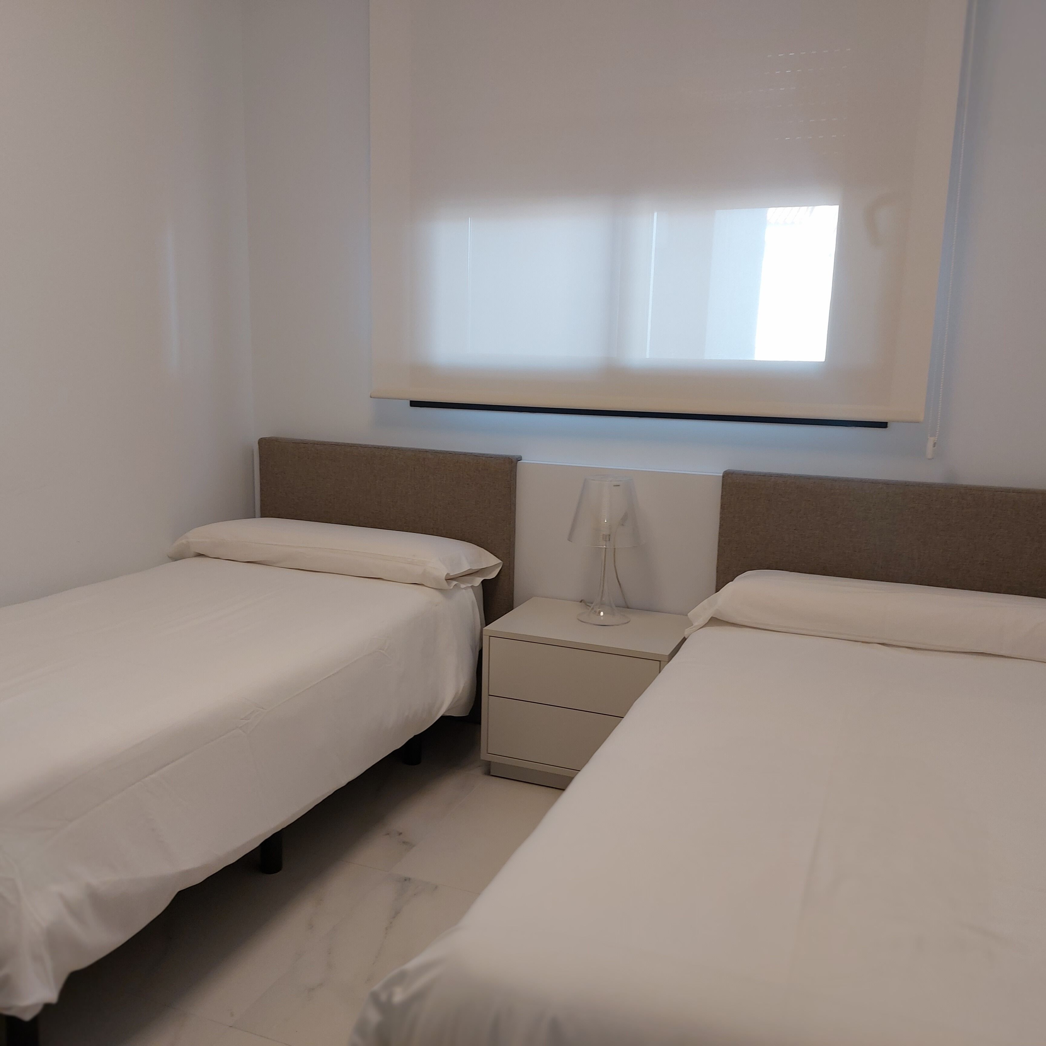 A bedroom with two beds, a nightstand, a lamp, and a window with blinds.