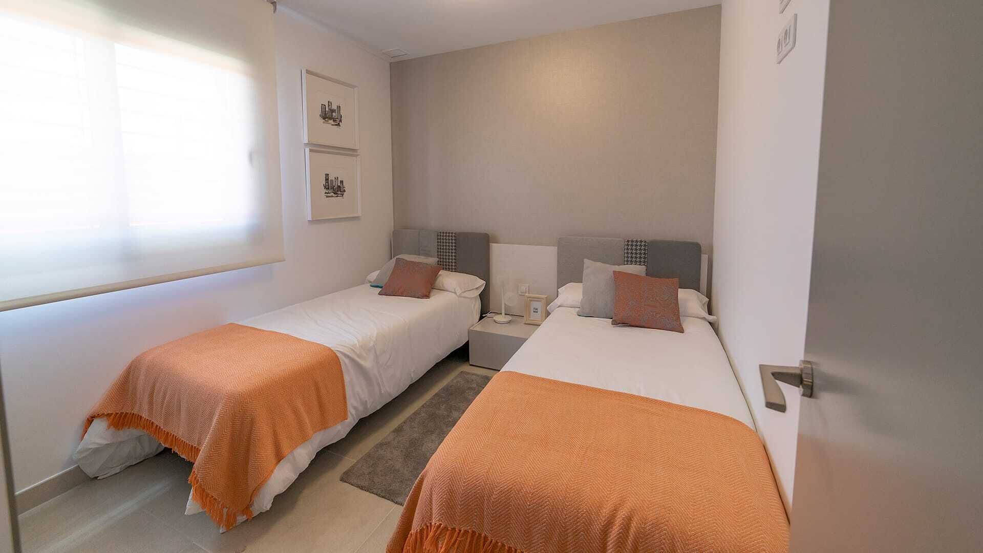 A hotel room with two single beds, a small bedside table with a lamp, a framed picture on the wall, and a window providing natural light.