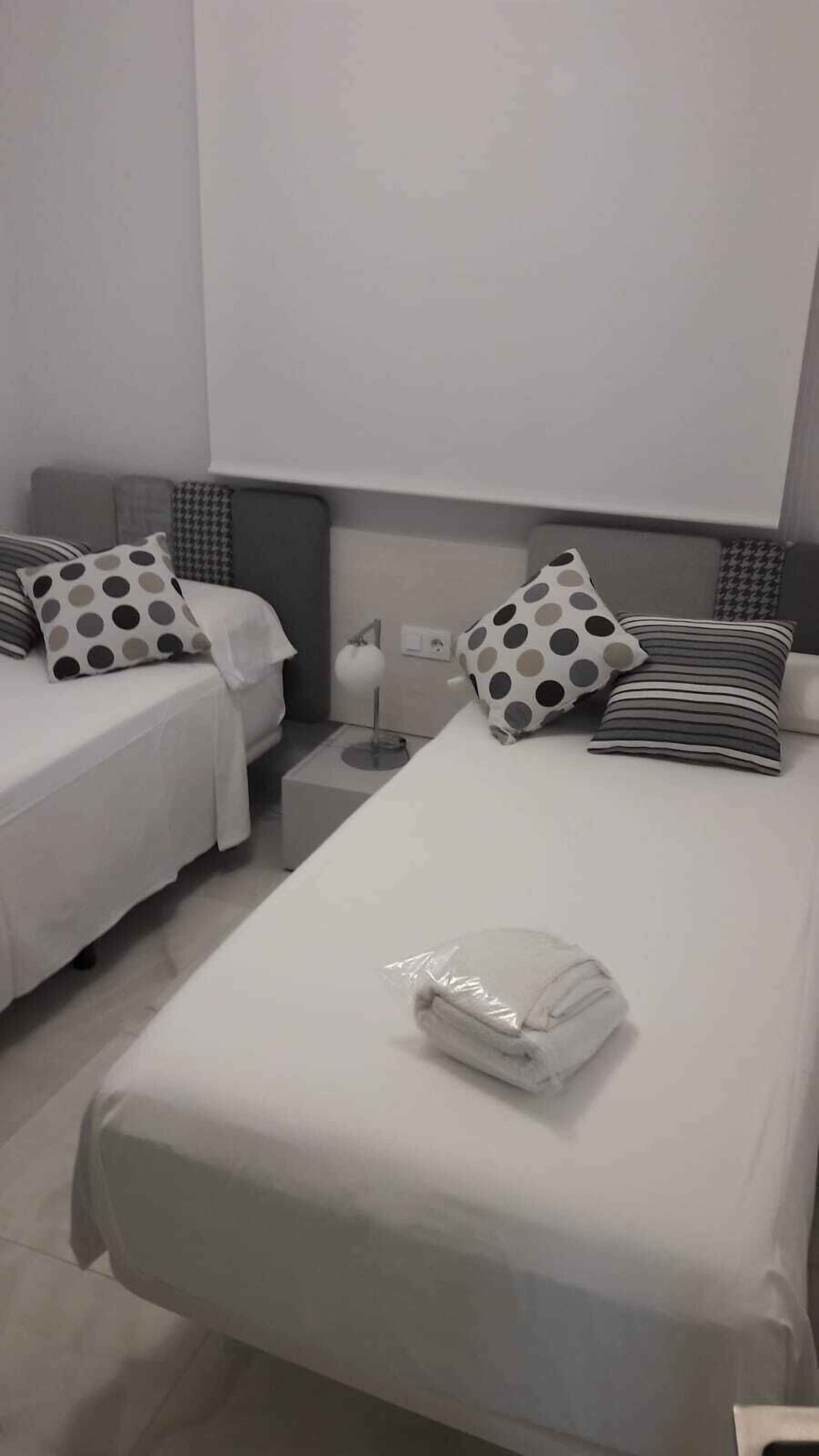 Een hotelkamer met twee eenpersoonsbedden, elk met een stippen kussen en een streepjeskussen. Er staat een klein nachtkastje met een lamp, en er ligt een gevouwen handdoek op het grotere bed.
