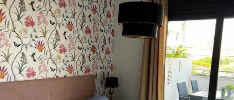 Habitación de hotel con una cama grande, papel tapiz floral, una lámpara en el techo y vistas al balcón exterior.
