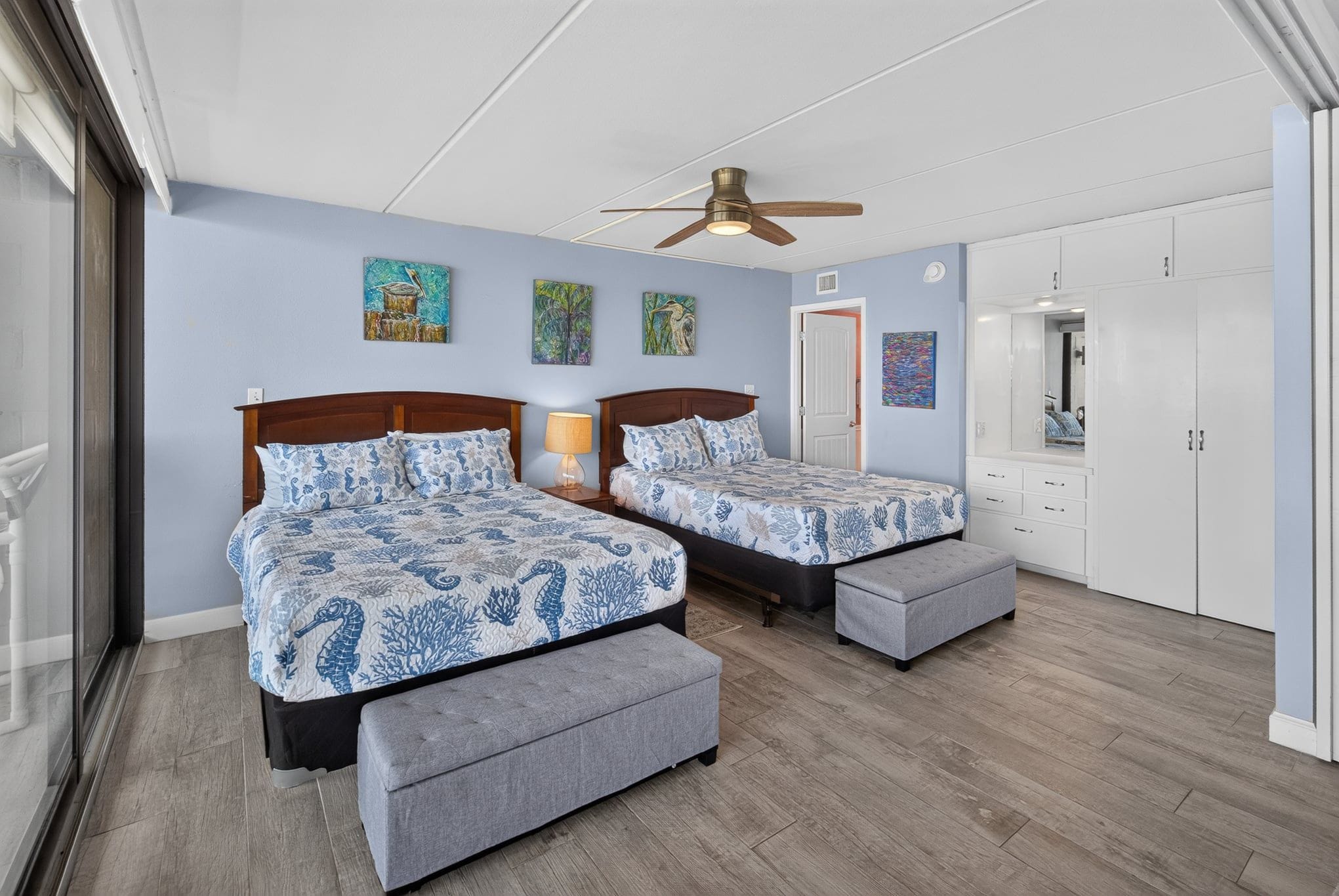 A bedroom with two beds, a ceiling fan, artwork on the walls, and a wardrobe.