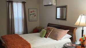 A neatly made bed with a white duvet and orange throw blanket, a bedside table with a lamp and a vase of flowers, and a wall-mounted air conditioner. - Poppies Place @ The Pleasant Street Suites in downtown Prescott (Prescott)
