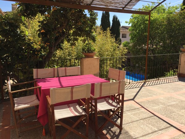 An outdoor patio with a table and chairs, surrounded by greenery and a view of a pool.