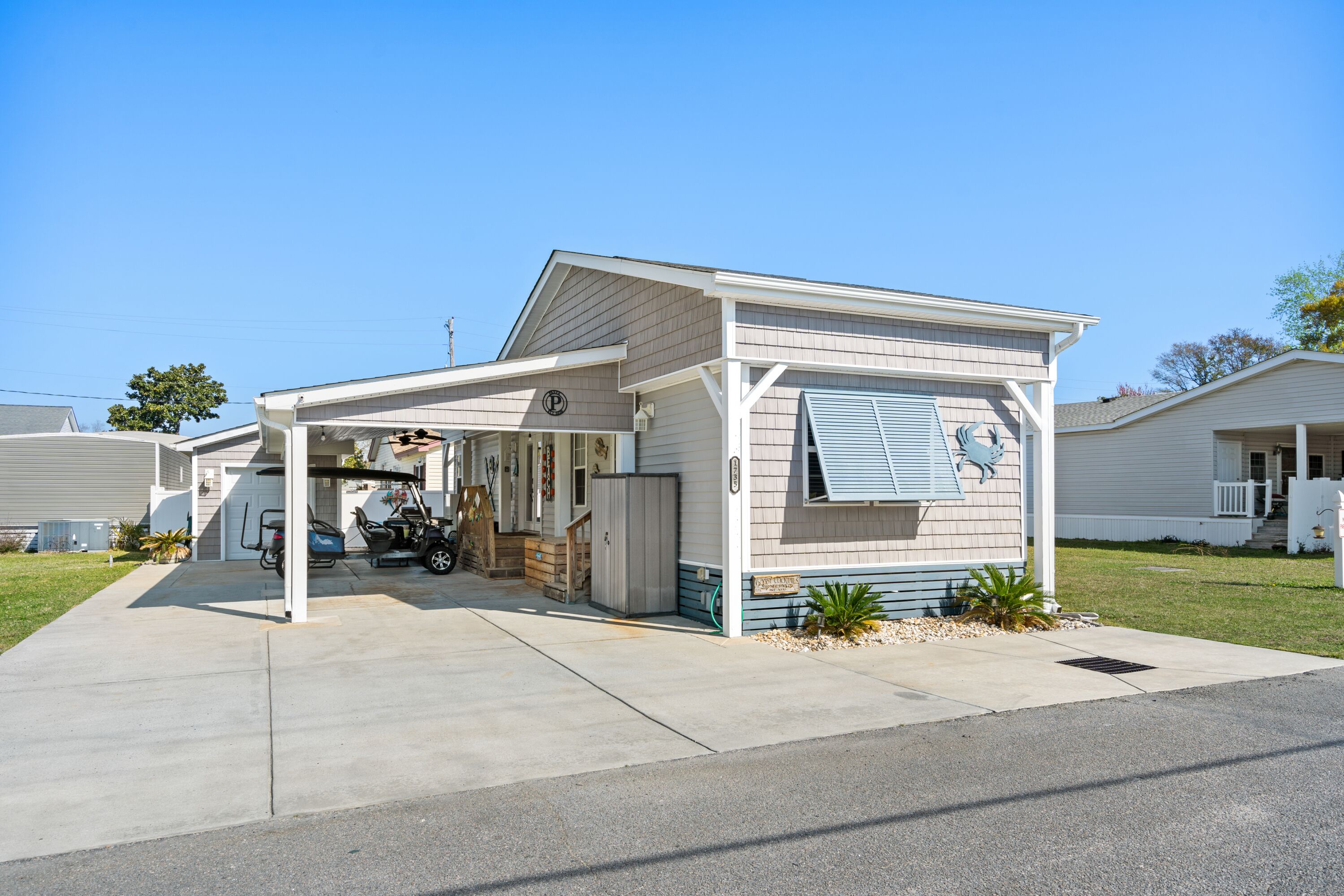 A single-story mobile home with a covered garage, a small porch, and a front yard with a palm plant.