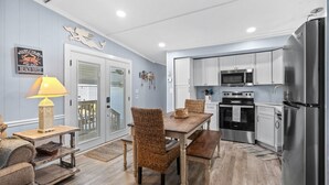 A kitchen with white cabinets, stainless steel appliances, and a dining area with wicker chairs and a wooden table. - Free Golf Cart, Beach Access/w Parking, + 3 pools! (Surfside Beach)