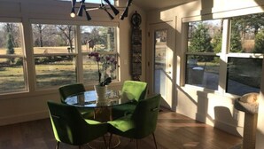 Een eethoek met een glazen tafel en groene stoelen, grote ramen met uitzicht op de omgeving, en een krabboom.