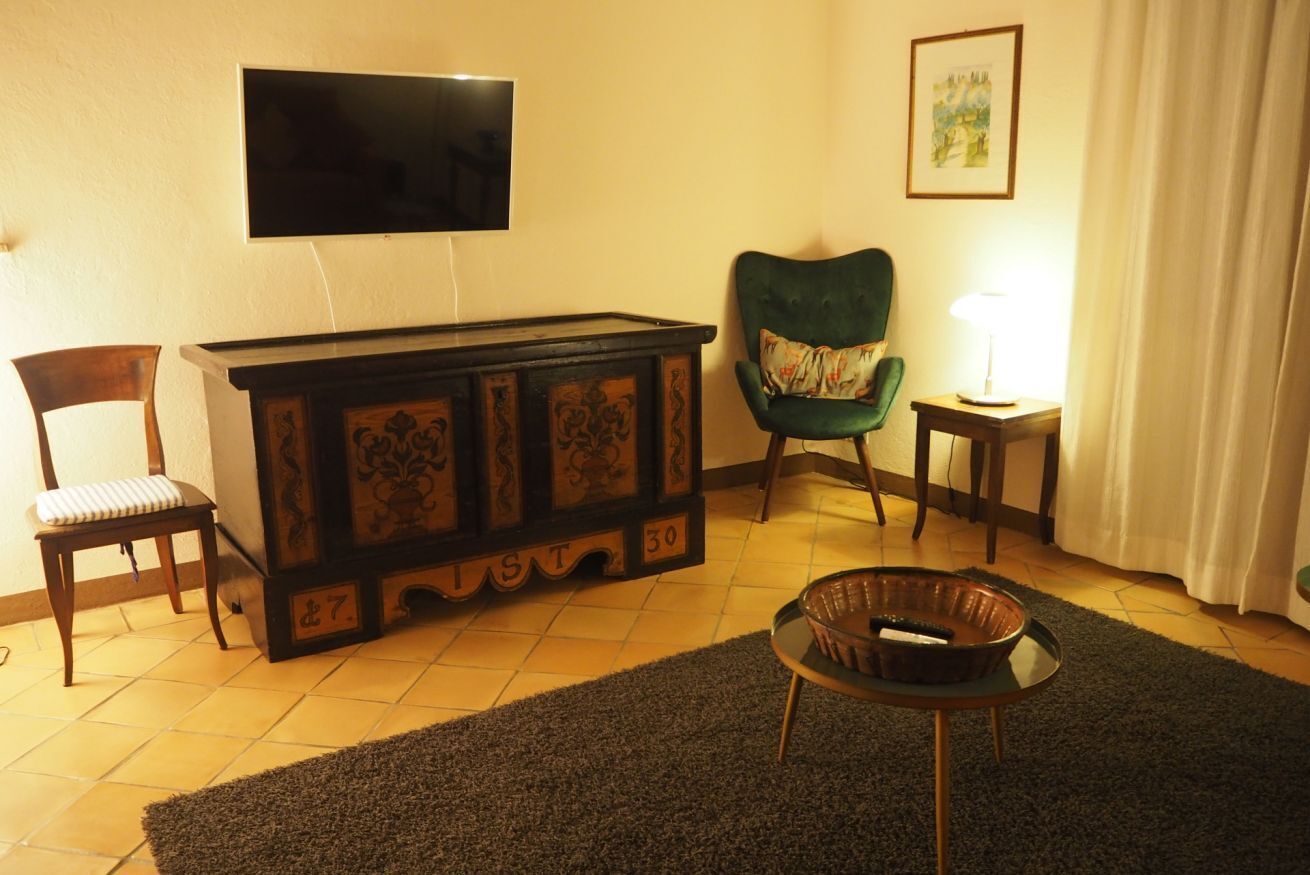 A living room with a television mounted on the wall, a green armchair, a wooden cabinet, a small table with a lamp, and a chair with a cushion.