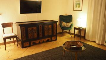 A living room with a television mounted on the wall, a green armchair, a wooden cabinet, a small table with a lamp, and a chair with a cushion.