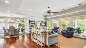 A spacious living room with hardwood floors, a dining area with wicker chairs, and a large window offering a view of greenery. - Beautiful Home w/ Private Pool in Sea Pines (Hilton Head Island)
