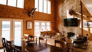 Un intérieur de chalet chaleureux avec une cheminée en pierre, des murs en bois, une grande télévision et un coin repas offrant une vue sur l’extérieur.
