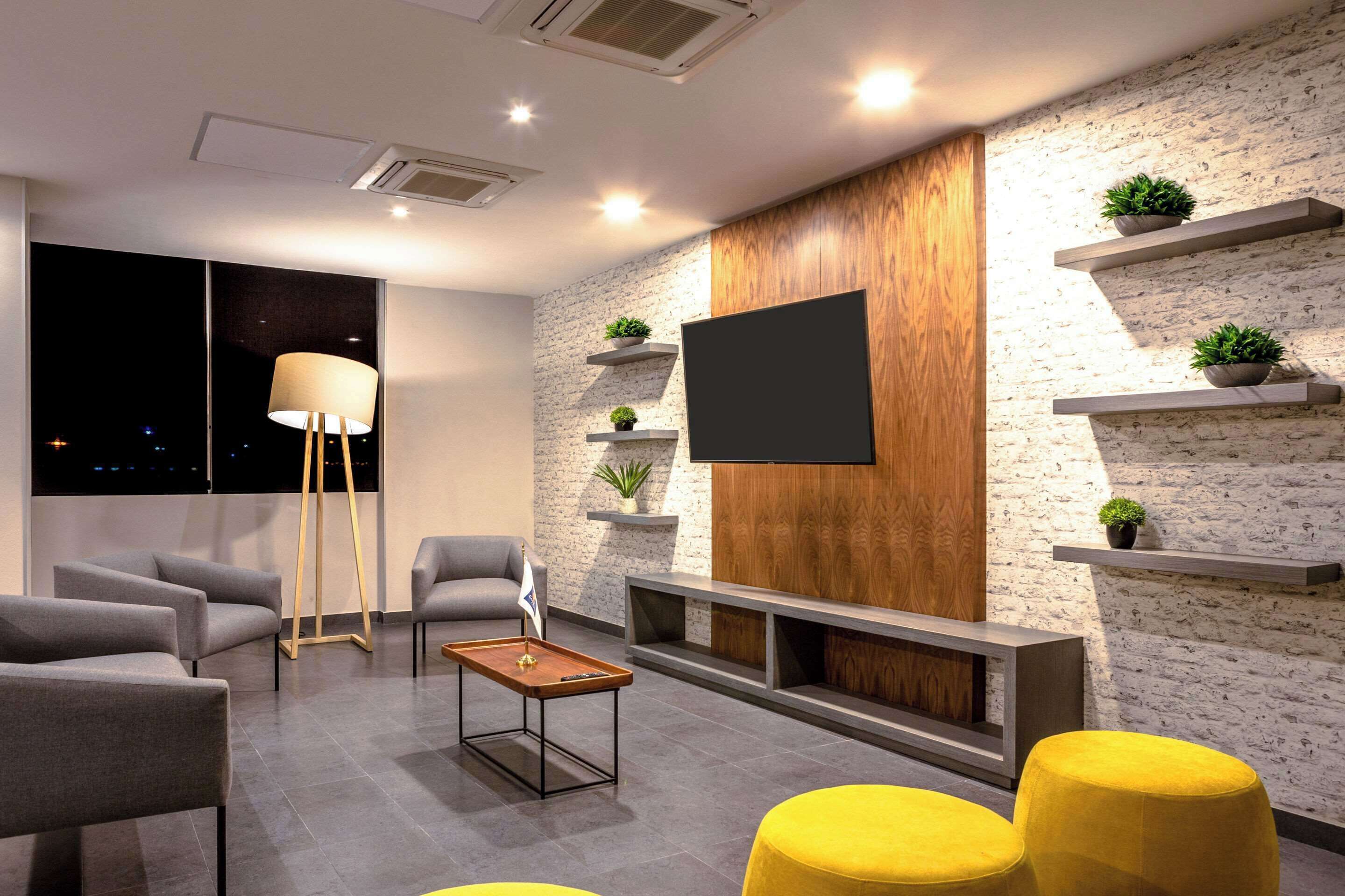 Een moderne woonkamer met grijze banken, een houten salontafel en gele poefs. Er zijn wandplanken met planten en een flatscreentelevisie.