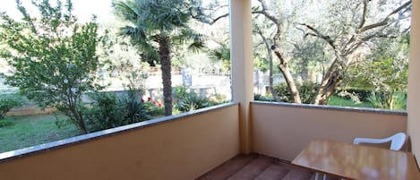 A balcony with a wooden table and white chairs, overlooking a garden with trees and a fence.