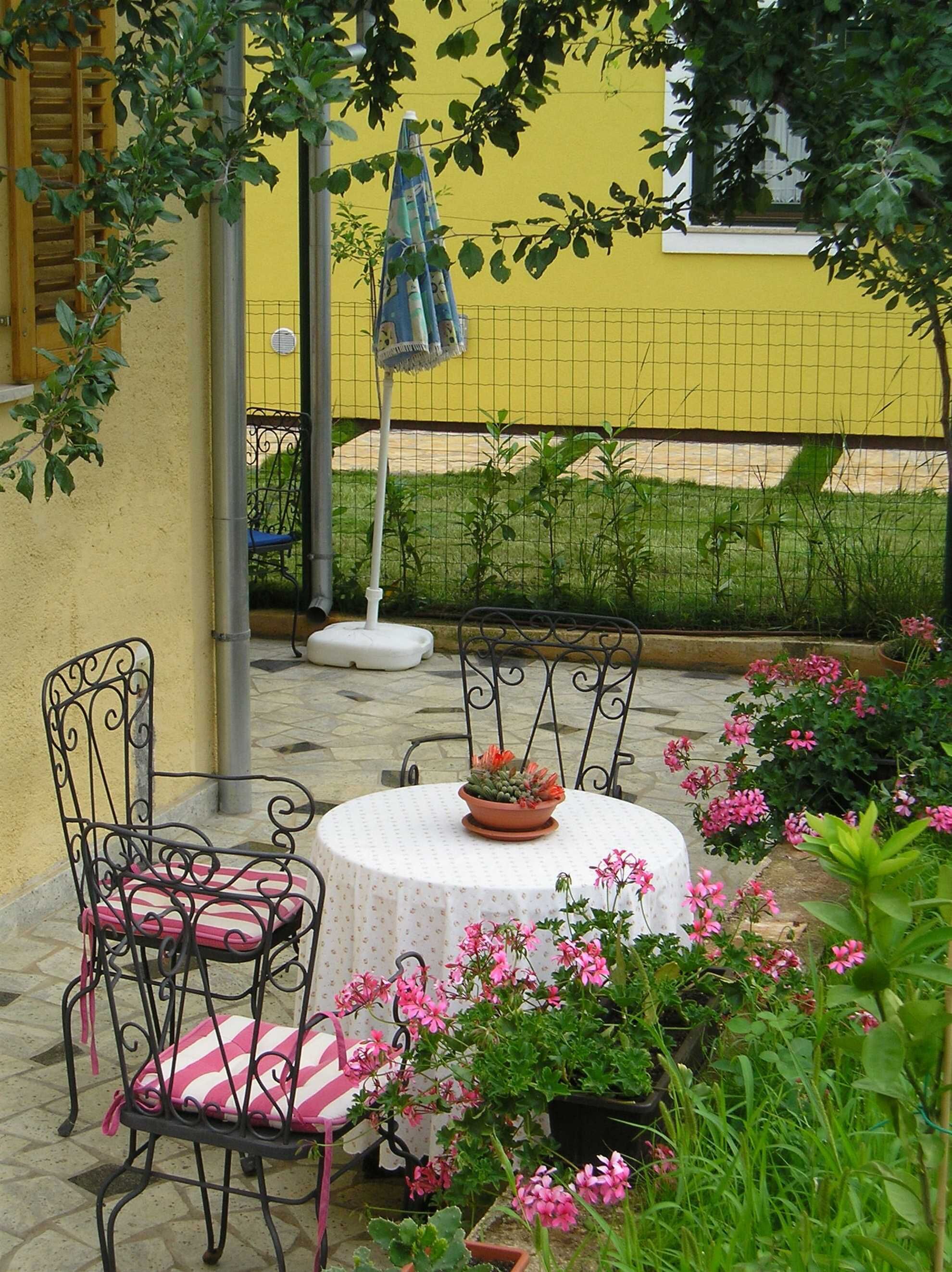 Een terras met twee metalen stoelen, een ronde tafel met een witte tafelkleed, een potplant en roze bloemen.