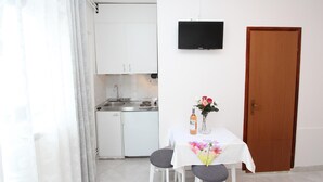 Ett kompakt kök med vita skåp, en inbyggd ugn och en diskho. Ett litet matbord med två stolar och en vas med blommor står framför en trädörr.
