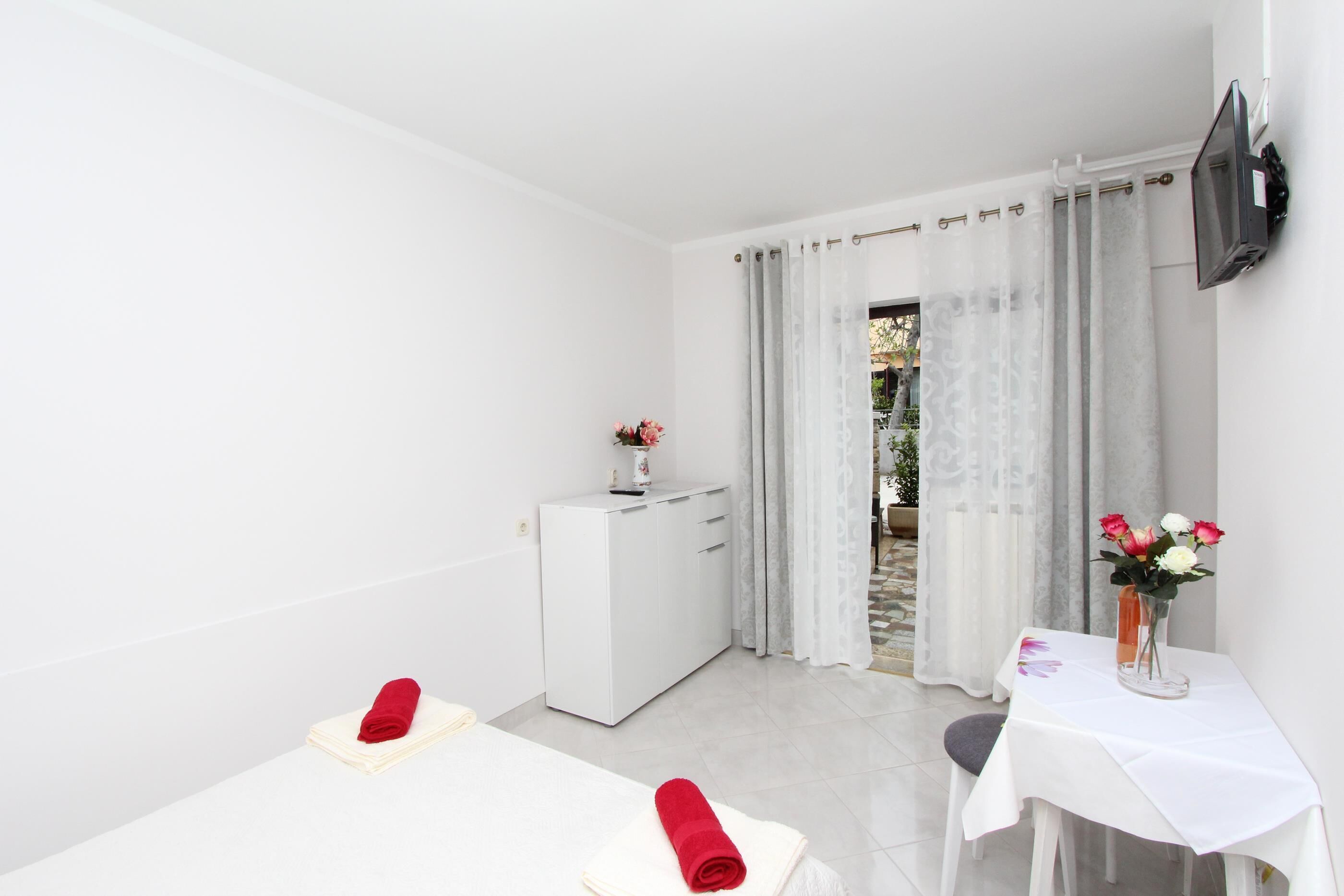 Ein helles, weißes Zimmer mit einem Bett, einem kleinen Tisch mit Blumenvase und einem weißen Schrank.