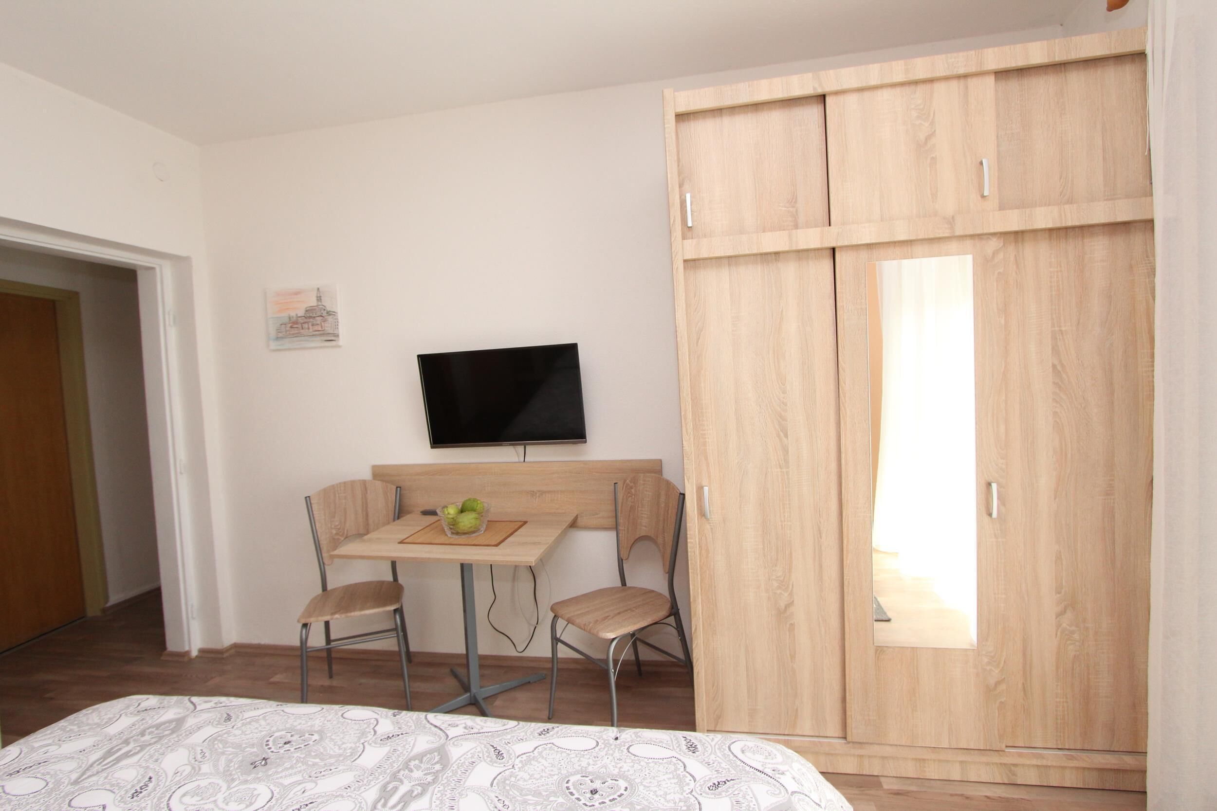 A bedroom with a bed, a wooden wardrobe, a TV on the wall, and a small table with chairs.