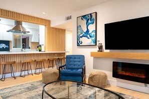 Una sala de estar moderna con un sillón azul, una mesa de centro de vidrio, una chimenea, una zona de cocina con detalles de madera y un televisor grande montado en la pared.