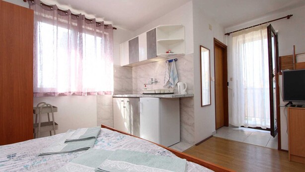 Quarto com cama, pequeno espaço de cozinha, televisão e banheiro.