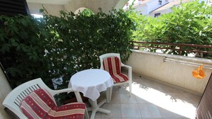 En lille udendørs terrasse med to hvide stole og et bord, omgivet af grønne planter.