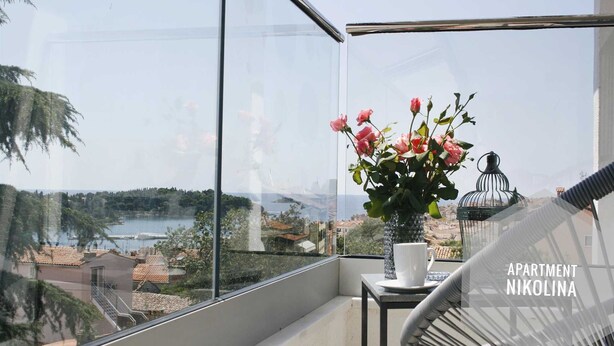 En balkon med frit udsyn over en sø og omkringliggende bygninger, en vase med lyserøde blomster, et lille bord med en kop, og en metalkonstruktion i form af et fuglebur.