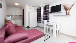 Un espace de vie compact avec un canapé rouge, une table à manger blanche, un téléviseur fixé au mur et un coin cuisine à l’arrière-plan.