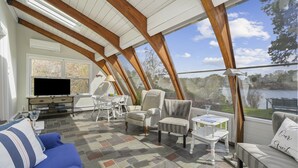 Une véranda avec vue sur un lac, comprenant un canapé, des fauteuils, une table et un téléviseur.