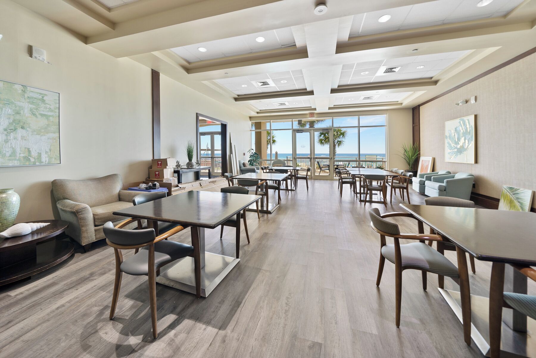 A spacious indoor area with multiple tables and chairs, a sofa, and a view of the ocean through large windows.