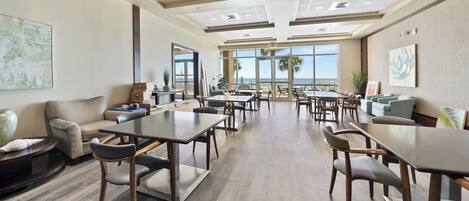 A spacious indoor area with multiple tables and chairs, a sofa, and a view of the ocean through large windows.